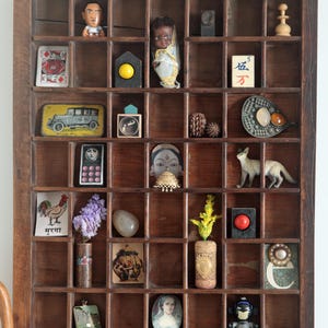 May include: A vintage wooden printer's tray displaying a collection of miniature items. The assortment includes a doll, playing card, toy car, figurines, a rooster illustration with text, a fox figurine, and a vase with flowers.