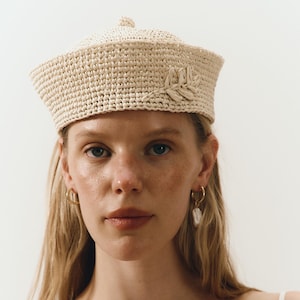 Puede incluir: Un sombrero tejido de color crema con un pequeño adorno floral decorativo. El sombrero tiene una copa redonda y un ala plana. La modelo lleva pendientes de aro dorados con un dije blanco en forma de pera. La modelo lleva un top blanco.