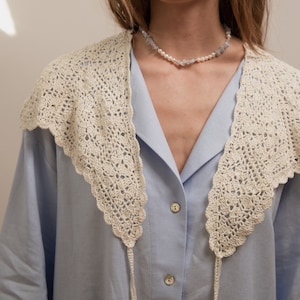 Peut inclure: Un col en crochet cr&egrave;me, &agrave; bord festonn&eacute;, est port&eacute; sur une chemise boutonn&eacute;e bleu clair. Un d&eacute;licat collier de perles et de pierres bleu clair orne le cou. Le col a de longues attaches fines.