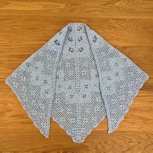May include: A light blue, handmade crochet shawl with a delicate openwork design. The shawl is triangular, with a scalloped edge and a floral pattern throughout. It is displayed on a wooden surface.