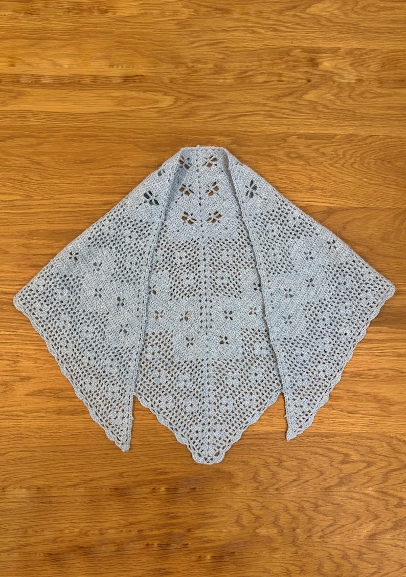 May include: A light blue, handmade crochet shawl with a delicate openwork design. The shawl is triangular, with a scalloped edge and a floral pattern throughout. It is displayed on a wooden surface.