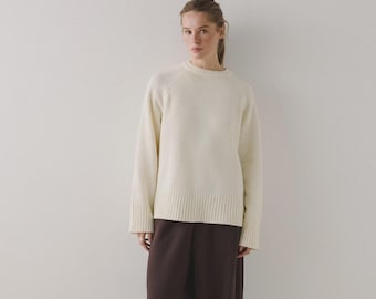 Könnte beinhalten: Ein cremefarbener Pullover mit langen Ärmeln und geripptem Saum und Bündchen. Der Pullover hat einen Rundhalsausschnitt und wird mit einer dunkelbraunen Hose kombiniert. Das Outfit ist schlicht und klassisch.