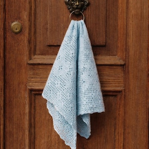 May include: A light blue crocheted cloth is draped over a brass door knocker. The cloth has a delicate openwork pattern and a scalloped edge. The door is made of wood with a warm, brown tone.