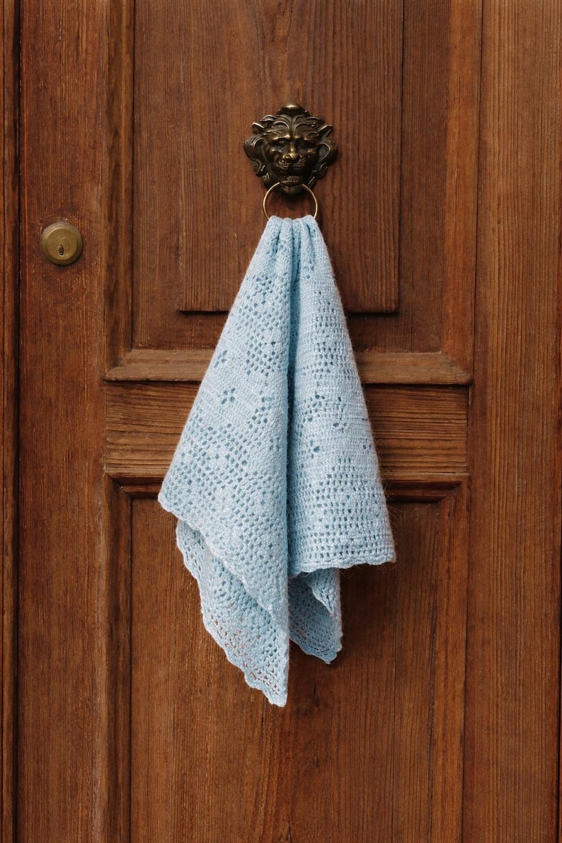 May include: A light blue crocheted cloth is draped over a brass door knocker. The cloth has a delicate openwork pattern and a scalloped edge. The door is made of wood with a warm, brown tone.