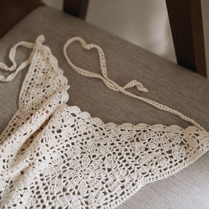 Peut inclure: Haut en crochet fait &agrave; la main de couleur cr&egrave;me, avec un bord festonn&eacute;. Le haut pr&eacute;sente un motif ajour&eacute; d&eacute;licat et des liens longs pour un ajustement r&eacute;glable. Le v&ecirc;tement est pr&eacute;sent&eacute; sur une chaise aux tons neutres.
