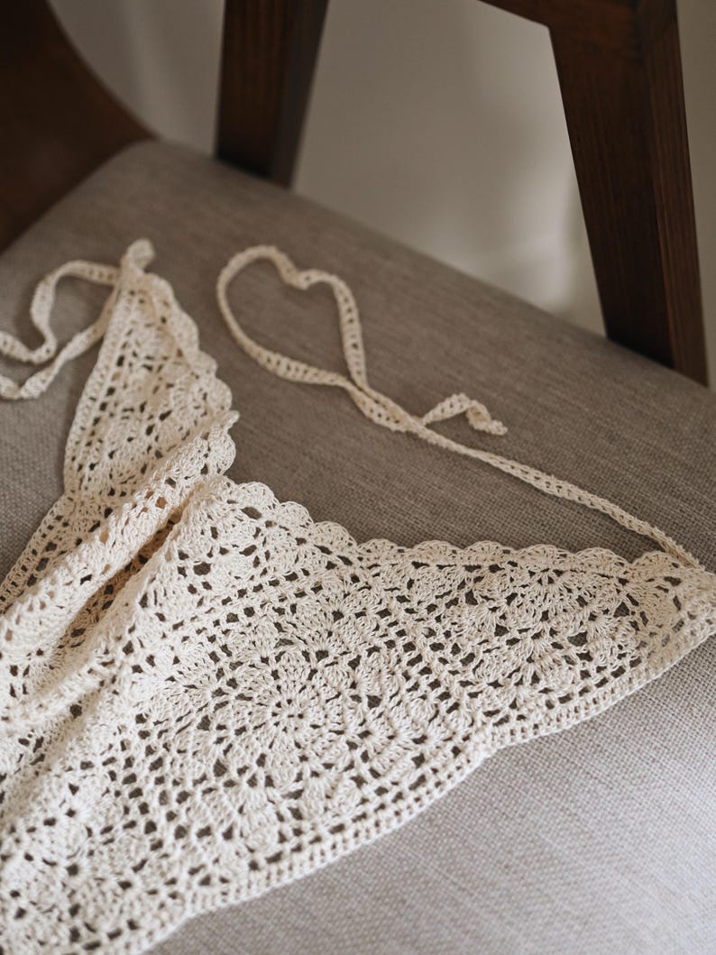 Peut inclure: Haut en crochet fait &agrave; la main de couleur cr&egrave;me, avec un bord festonn&eacute;. Le haut pr&eacute;sente un motif ajour&eacute; d&eacute;licat et des liens longs pour un ajustement r&eacute;glable. Le v&ecirc;tement est pr&eacute;sent&eacute; sur une chaise aux tons neutres.