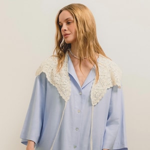 Peut inclure: Une chemise bleu clair avec un col en crochet cr&egrave;me. Le col pr&eacute;sente un motif ajour&eacute; d&eacute;licat. La chemise a des manches longues et une coupe ample. Une ficelle blanche est attach&eacute;e au col.