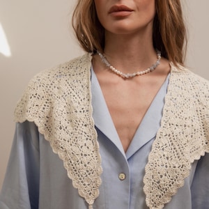 Peut inclure: Un col en crochet cr&egrave;me, avec un bord festonn&eacute;, est port&eacute; sur une chemise boutonn&eacute;e bleu clair. Un d&eacute;licat collier de perles et de pierres bleu clair orne le cou. Le col a de longues attaches fines.