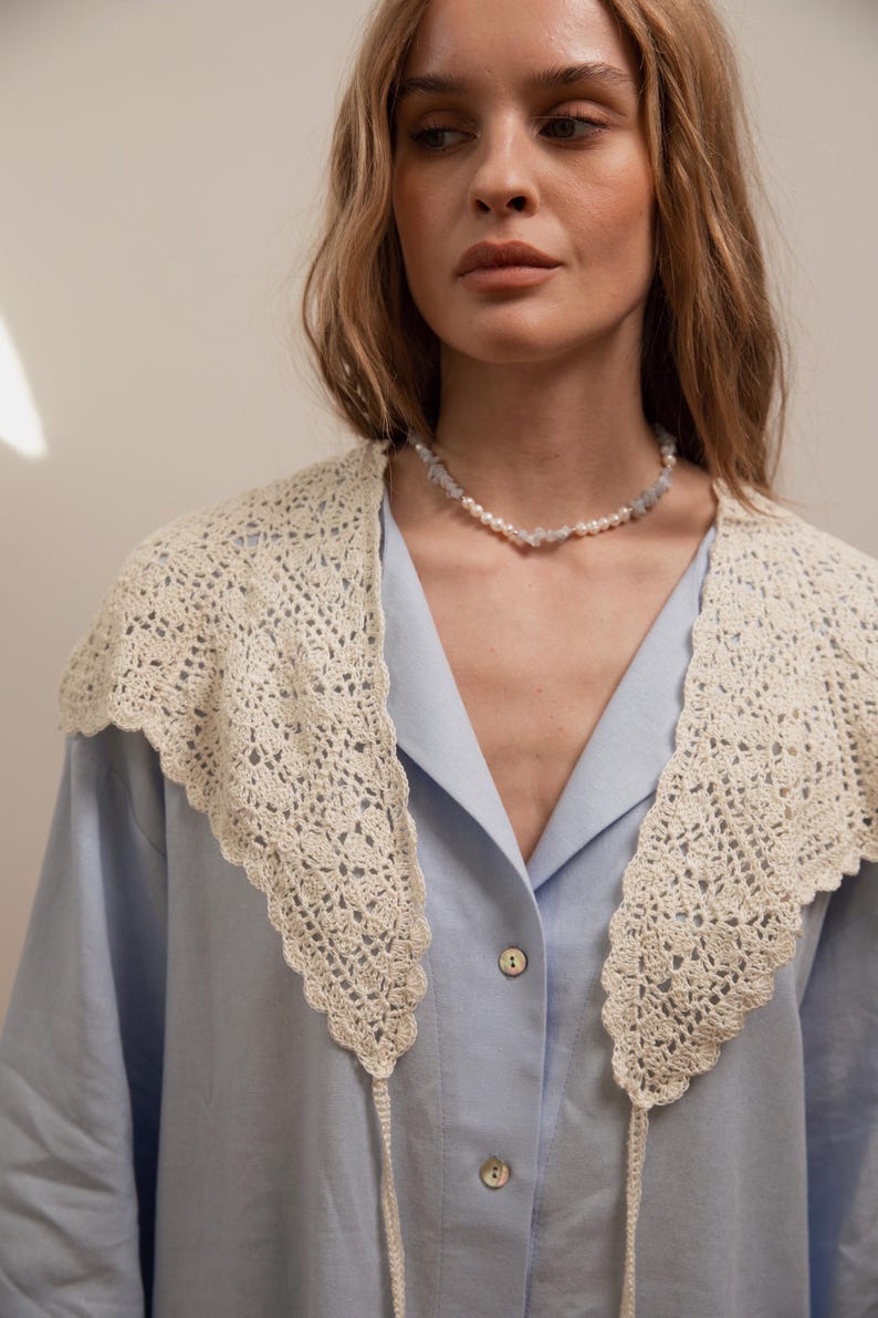 Peut inclure: Un col en crochet cr&egrave;me, avec un bord festonn&eacute;, est port&eacute; sur une chemise boutonn&eacute;e bleu clair. Un d&eacute;licat collier de perles et de pierres bleu clair orne le cou. Le col a de longues attaches fines.