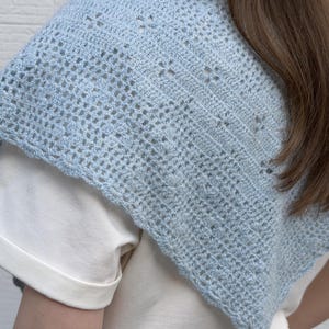 May include: A light blue crochet shawl with an open, airy design. The shawl is worn over a white shirt, showcasing the delicate, handmade texture. The crochet pattern features small, evenly spaced holes, creating a lightweight and breathable accessory.