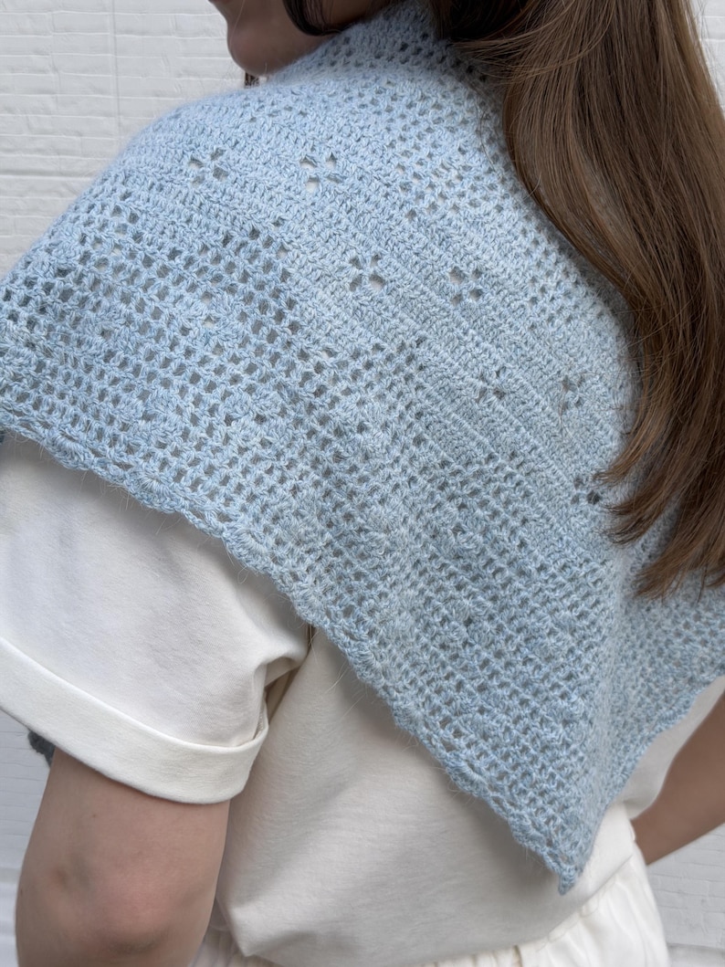 May include: A light blue crochet shawl with an open, airy design. The shawl is worn over a white shirt, showcasing the delicate, handmade texture. The crochet pattern features small, evenly spaced holes, creating a lightweight and breathable accessory.