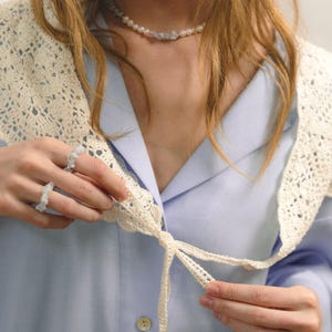 Peut inclure: Une chemise bleu clair avec un col en crochet cr&egrave;me et une cravate assortie. Un collier de perles et de pierres et une bague en pierre sont &eacute;galement visibles. Le col est nou&eacute; en n&oelig;ud.