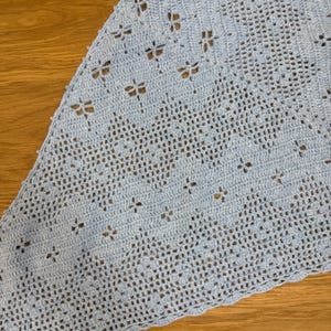 May include: A light blue, triangular crochet shawl. The shawl features a delicate openwork design with small, flower-like cutouts. The edges have a scalloped trim. The shawl is made of soft yarn, perfect for a light layer.