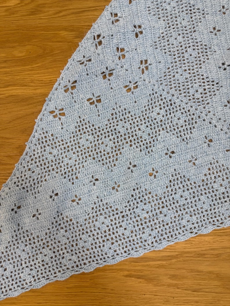 May include: A light blue, triangular crochet shawl. The shawl features a delicate openwork design with small, flower-like cutouts. The edges have a scalloped trim. The shawl is made of soft yarn, perfect for a light layer.