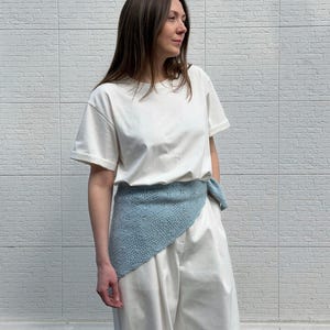 May include: A woman wearing a white t-shirt and white pants, with a light blue knitted wrap tied around the waist. The wrap has a delicate, openwork design. The background is a white brick wall.