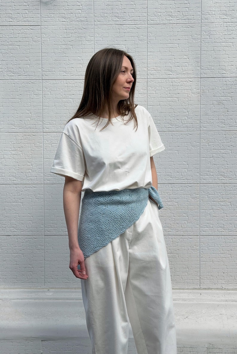 May include: A woman wearing a white t-shirt and white pants, with a light blue knitted wrap tied around the waist. The wrap has a delicate, openwork design. The background is a white brick wall.