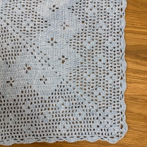 May include: A light blue crocheted blanket with a delicate openwork pattern. The blanket features a scalloped edge and is displayed on a wooden surface. The crochet design includes small floral motifs.