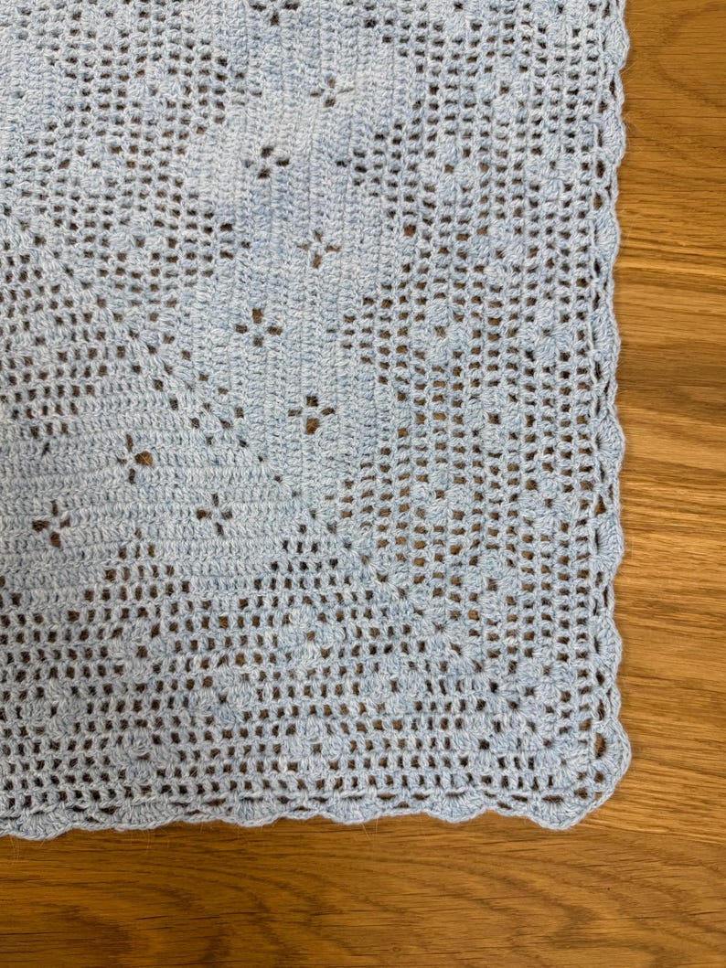 May include: A light blue crocheted blanket with a delicate openwork pattern. The blanket features a scalloped edge and is displayed on a wooden surface. The crochet design includes small floral motifs.