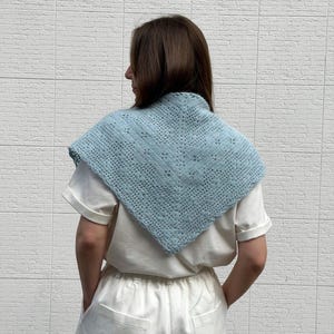 May include: A light blue crocheted shawl with a geometric openwork design. The shawl is worn over a white t-shirt. The shawl is a square shape with a pointed bottom edge. The background is a white brick wall.