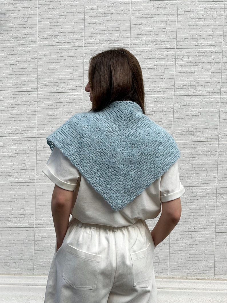 May include: A light blue crocheted shawl with a geometric openwork design. The shawl is worn over a white t-shirt. The shawl is a square shape with a pointed bottom edge. The background is a white brick wall.