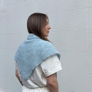 May include: A light blue knitted shawl with a delicate openwork pattern. The shawl is triangular in shape and worn over the shoulders. The garment is made of a soft, possibly wool, material.