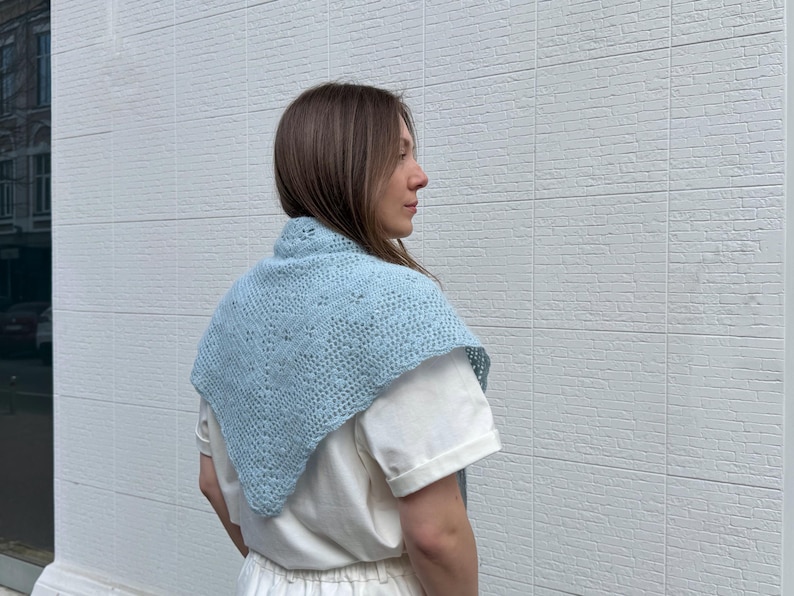 May include: A light blue knitted shawl with a delicate openwork pattern. The shawl is triangular in shape and worn over the shoulders. The garment is made of a soft, possibly wool, material.