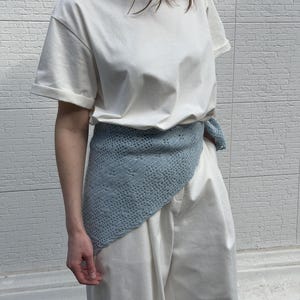 May include: A light blue crocheted wrap worn over a white t-shirt and white pants. The wrap is draped around the waist. The background is a white brick wall.