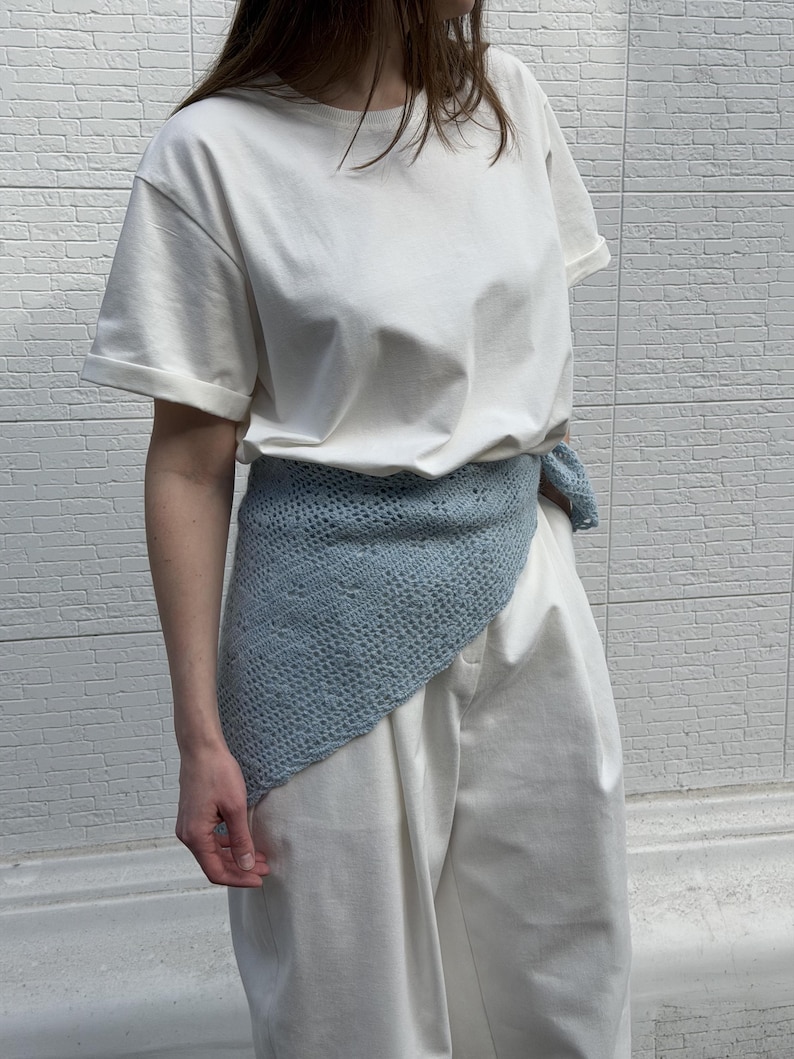 May include: A light blue crocheted wrap worn over a white t-shirt and white pants. The wrap is draped around the waist. The background is a white brick wall.