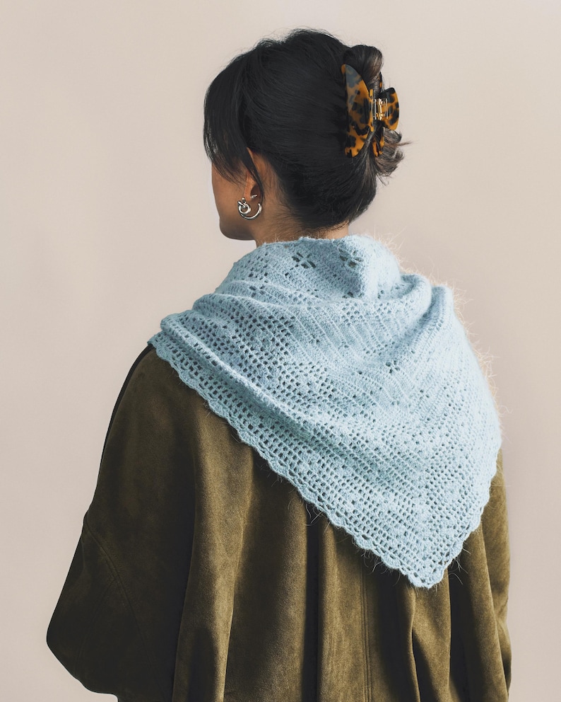 May include: A light blue, crocheted shawl with a delicate openwork pattern. The shawl is draped over a dark green jacket. A tortoiseshell hair clip is visible in the person's dark hair. Silver hoop earrings complete the look. The shawl's texture and color create a soft, feminine aesthetic.