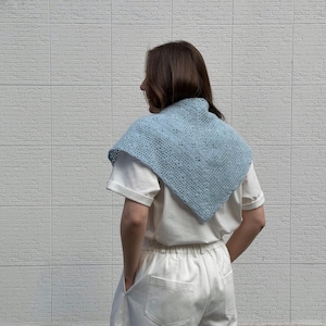 May include: A light blue crocheted shawl with a square shape and a delicate openwork pattern. The shawl is worn over a white t-shirt and paired with white pants. The background is a white brick wall.