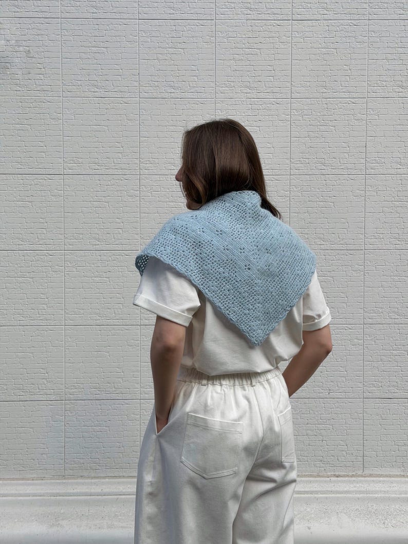May include: A light blue crocheted shawl with a square shape and a delicate openwork pattern. The shawl is worn over a white t-shirt and paired with white pants. The background is a white brick wall.