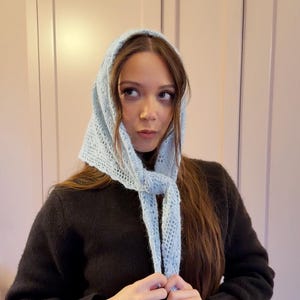May include: A light blue, open-weave knitted headscarf with a tie closure. The head covering is worn over the head and tied under the chin. The scarf is made of a soft, light blue yarn.