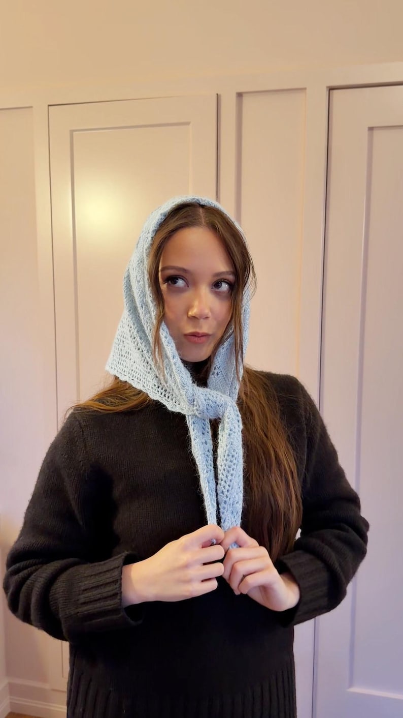 May include: A light blue, open-weave knitted headscarf with a tie closure. The head covering is worn over the head and tied under the chin. The scarf is made of a soft, light blue yarn.
