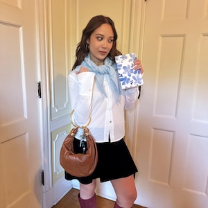 May include: A brown leather handbag with gold-tone handles. The bag is paired with a white button-down shirt, a black skirt, and burgundy knee-high boots. A light blue scarf and a floral-patterned notebook complete the ensemble.