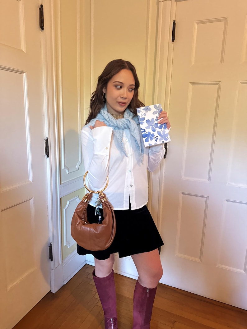 May include: A brown leather handbag with gold-tone handles. The bag is paired with a white button-down shirt, a black skirt, and burgundy knee-high boots. A light blue scarf and a floral-patterned notebook complete the ensemble.