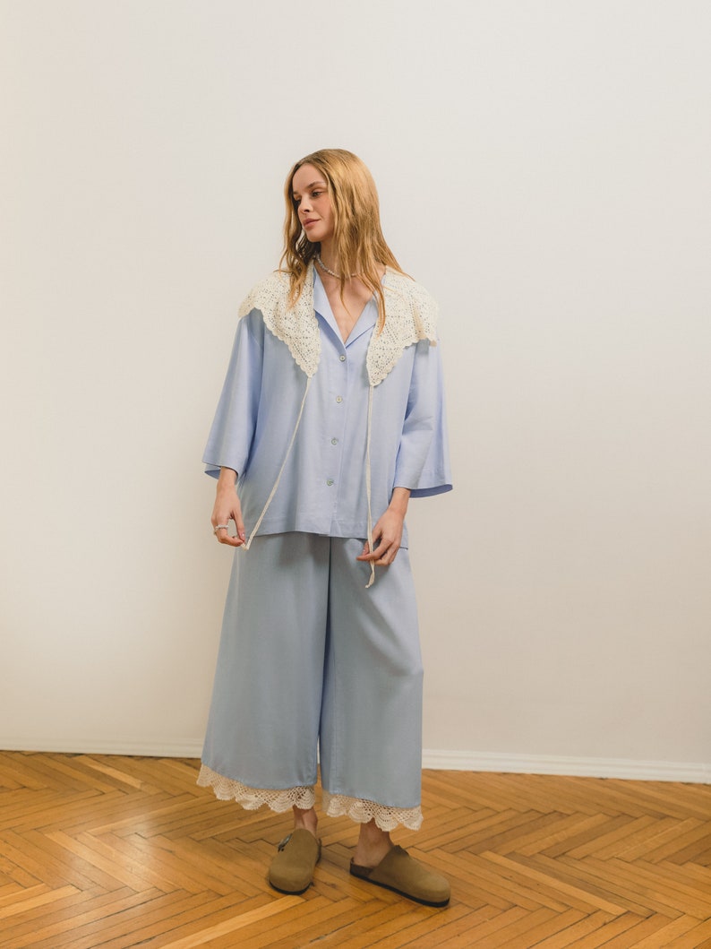 Peut inclure: Un ensemble de pyjama bleu clair comprenant une chemise boutonn&eacute;e avec un col en crochet et un pantalon large avec une bordure en dentelle. L'ensemble est associ&eacute; &agrave; des sabots marron. L'ensemble est fait d'un tissu doux et l&eacute;ger.