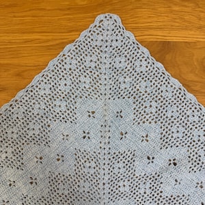 May include: A light blue, crocheted shawl with a delicate openwork pattern. The triangular shawl features a scalloped edge and a repeating floral design. The handmade textile is displayed on a wooden surface.