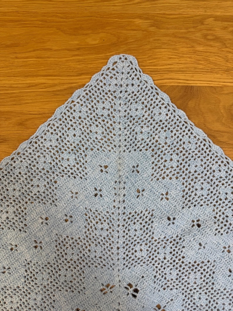 May include: A light blue, crocheted shawl with a delicate openwork pattern. The triangular shawl features a scalloped edge and a repeating floral design. The handmade textile is displayed on a wooden surface.