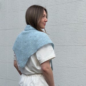 May include: A light blue, crocheted shawl with a delicate openwork pattern. The shawl is triangular in shape and worn over the shoulders. The person in the image is wearing a white t-shirt and white pants.