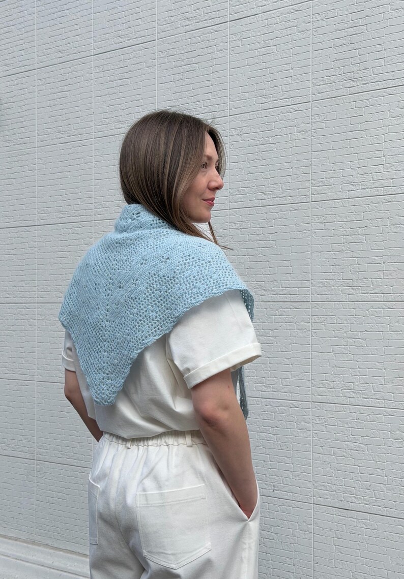 May include: A light blue, crocheted shawl with a delicate openwork pattern. The shawl is triangular in shape and worn over the shoulders. The person in the image is wearing a white t-shirt and white pants.