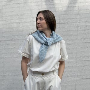 May include: A light blue, open-weave scarf is worn over a white t-shirt and white pants. The scarf is knotted at the front. The background is a white brick wall. The outfit is casual and comfortable.