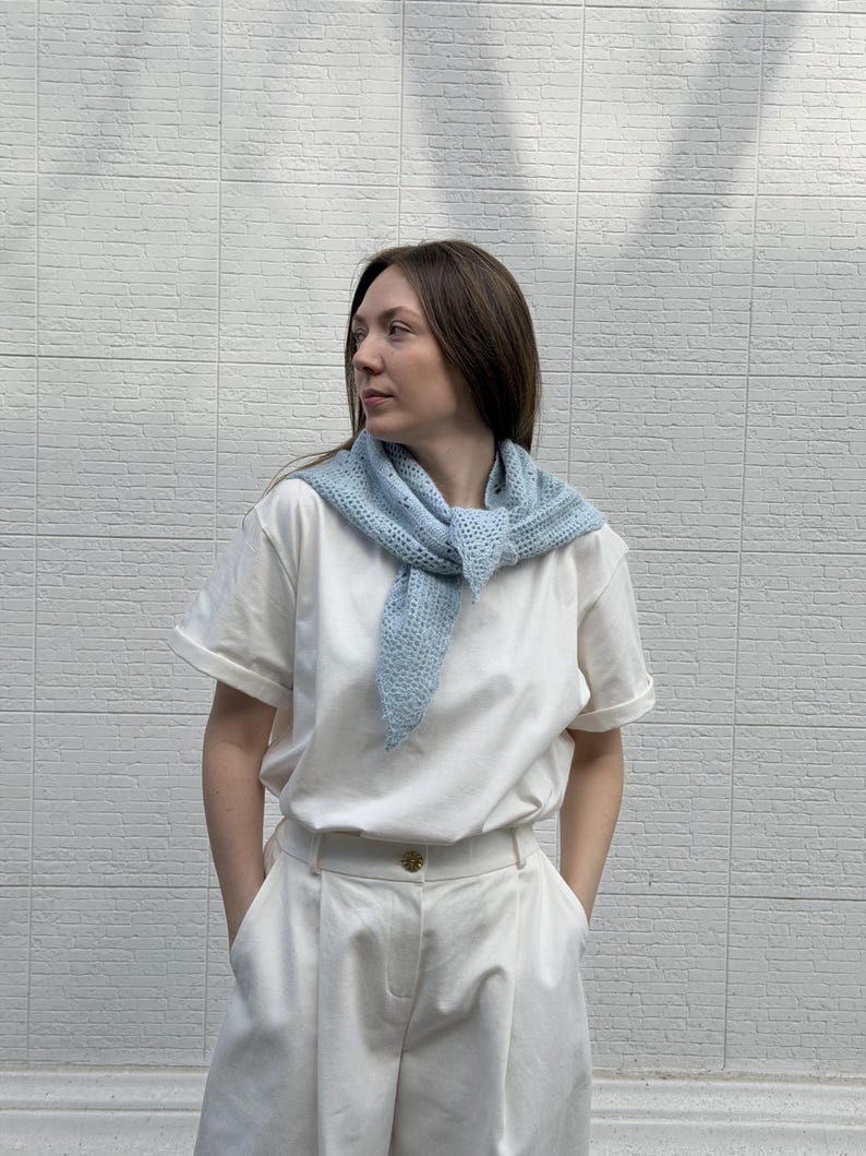 May include: A light blue, open-weave scarf is worn over a white t-shirt and white pants. The scarf is knotted at the front. The background is a white brick wall. The outfit is casual and comfortable.