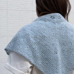 May include: A light blue crocheted shawl with a delicate openwork design. The shawl is draped over a white shirt, showcasing its intricate pattern and soft color. The shawl's texture and color create a cozy and stylish accessory.