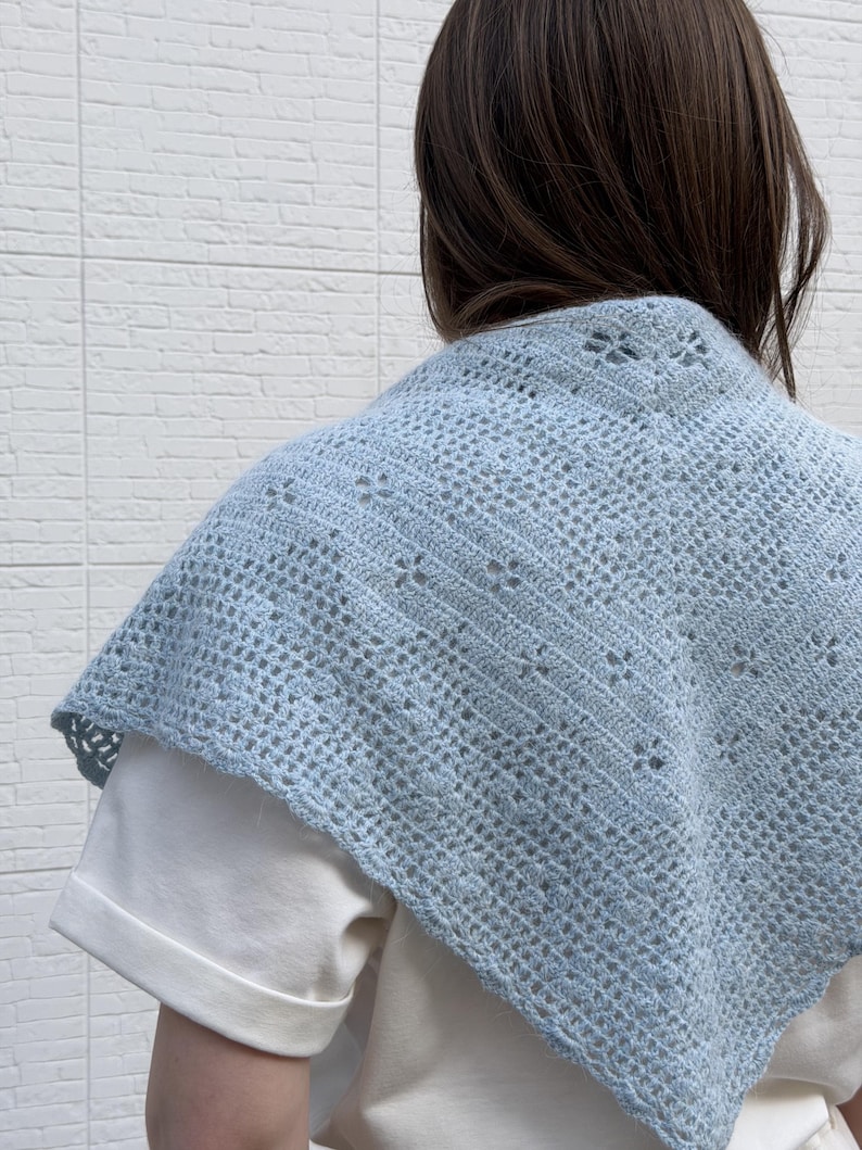 May include: A light blue crocheted shawl with a delicate openwork design. The shawl is draped over a white shirt, showcasing its intricate pattern and soft color. The shawl's texture and color create a cozy and stylish accessory.