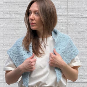 May include: A light blue crocheted shawl with a delicate openwork design. The shawl is worn over a white t-shirt and is held by the wearer's hands. The background is a white brick wall.