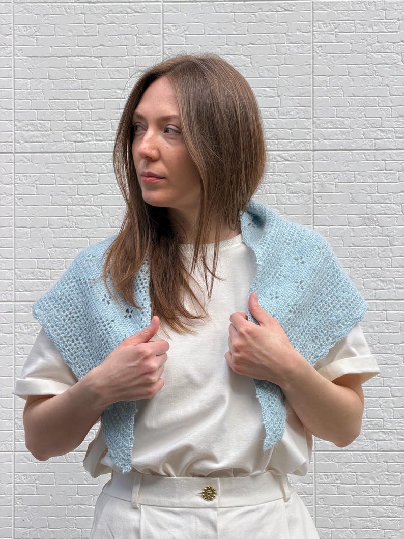May include: A light blue crocheted shawl with a delicate openwork design. The shawl is worn over a white t-shirt and is held by the wearer's hands. The background is a white brick wall.
