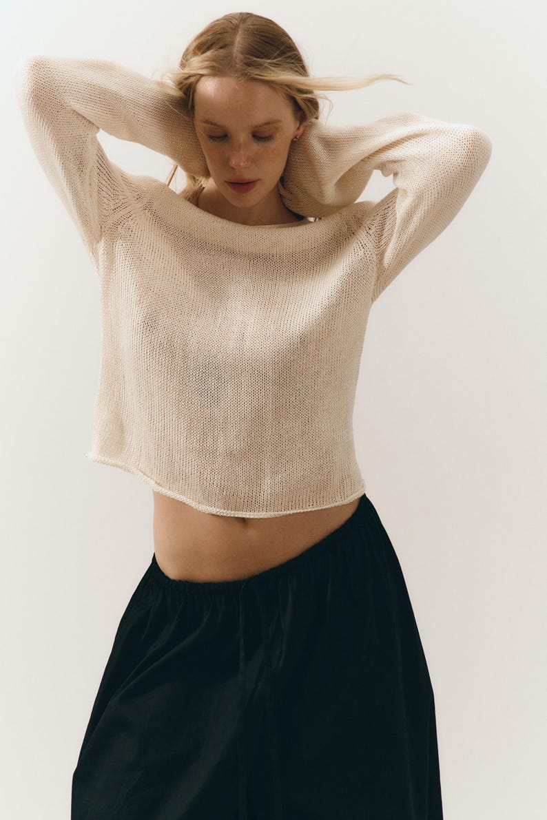 Puede incluir: Un su&eacute;ter de punto crema con tejido abierto, mangas largas y cuello barco. El su&eacute;ter se combina con pantalones negros de pernera ancha. Los brazos del modelo est&aacute;n levantados y el fondo es blanco. La textura del su&eacute;ter es visible.