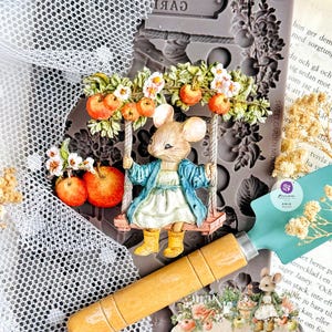 May include: A whimsical scene featuring a mouse on a swing adorned with apples and flowers. A gardening trowel and a mold with the word "WELCOME" are also visible. The overall theme suggests a garden or cottagecore aesthetic.