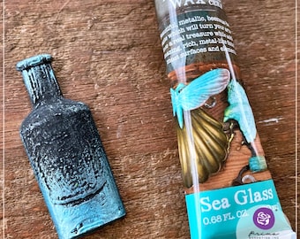 May include: A small glass bottle with a teal and black metallic finish. The bottle is sitting on a wooden surface. A tube of art alchemy metallic wax in the color sea glass is also shown. The tube has a label with the text "Sea Glass" and "0.68 FL. OZ."
