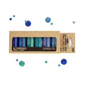 May include: A glitter set in a cardboard box labeled "Caribbean Sea." The set includes six glitter tubes in shades of blue and green. Loose glitter dots in matching colors are scattered around the box. The text "Glitter Set" is also visible.
